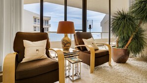 Un salon moderne avec deux fauteuils marron, une table d’appoint avec une lampe, une plante en pot et une vue sur un immeuble à travers de grandes fenêtres.
