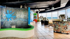 Ein moderner Poolbereich im Innenbereich mit einer Wand aus Mosaiken mit der Aufschrift „SPLASH“, umgeben von bunten Sitzgelegenheiten und großen Fenstern mit Blick auf den Ozean.