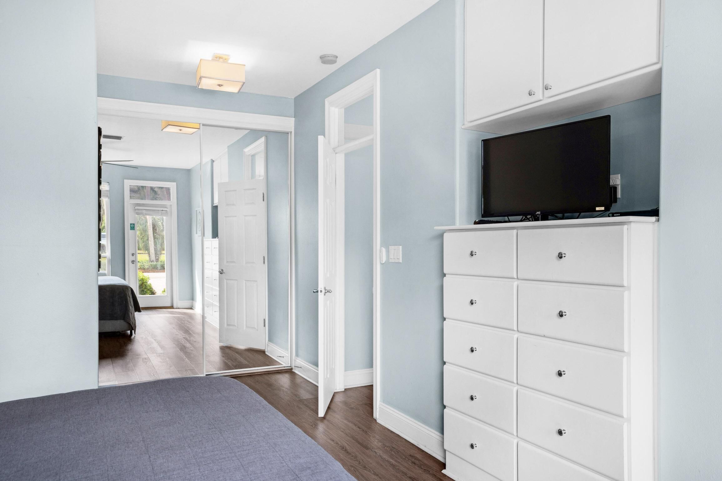 Una habitación de hotel con cama, cómoda blanca, televisor y puerta que da a otra habitación.