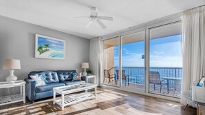 A spacious living area with a blue leather sofa, a coffee table, and a large sliding glass door leading to a balcony with a view of the ocean. - Majestic Beach Towers 1-1210 (Panama City Beach)