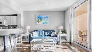 A modern living room with a blue sofa, white coffee table, and a painting of a beach scene. - Majestic Beach Towers 1-1210 (Panama City Beach)