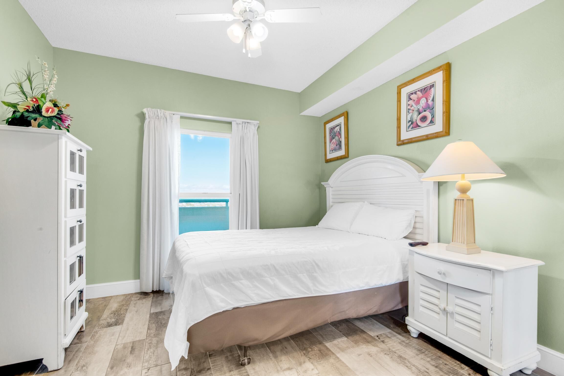 Une chambre à coucher comprenant un lit, une table de chevet, une commode, un ventilateur de plafond et une fenêtre avec des rideaux.