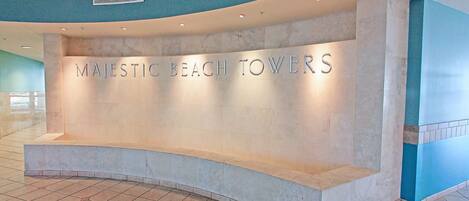 A reception desk with the sign "MAJESTIC BEACH TOWERS" in a modern lobby with tiled flooring and curved ceiling design.