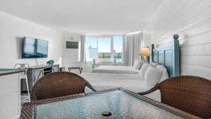 A hotel room with a bed, a TV, a dining table, wicker chairs, and a window with curtains. - Top of the Gulf 807 (Panama City Beach)