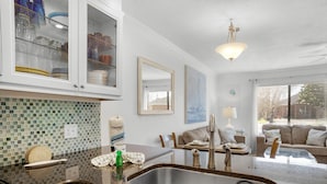 A modern kitchen with a granite countertop, stainless steel sink, and white cabinets. There is a dining area with a sofa and a coffee table. - Sandprints B3 - Miramar Sands (Miramar Beach)