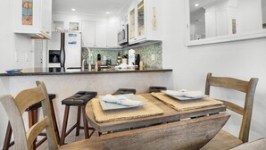 A modern kitchen with white cabinets, a stainless steel refrigerator, and a tiled backsplash. There is a wooden dining table with two chairs and a towel on it. - Sandprints B3 - Miramar Sands (Miramar Beach)