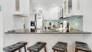 A modern kitchen with white cabinets, a tiled backsplash, and a bar area with stools. - Sandprints B3 - Miramar Sands (Miramar Beach)