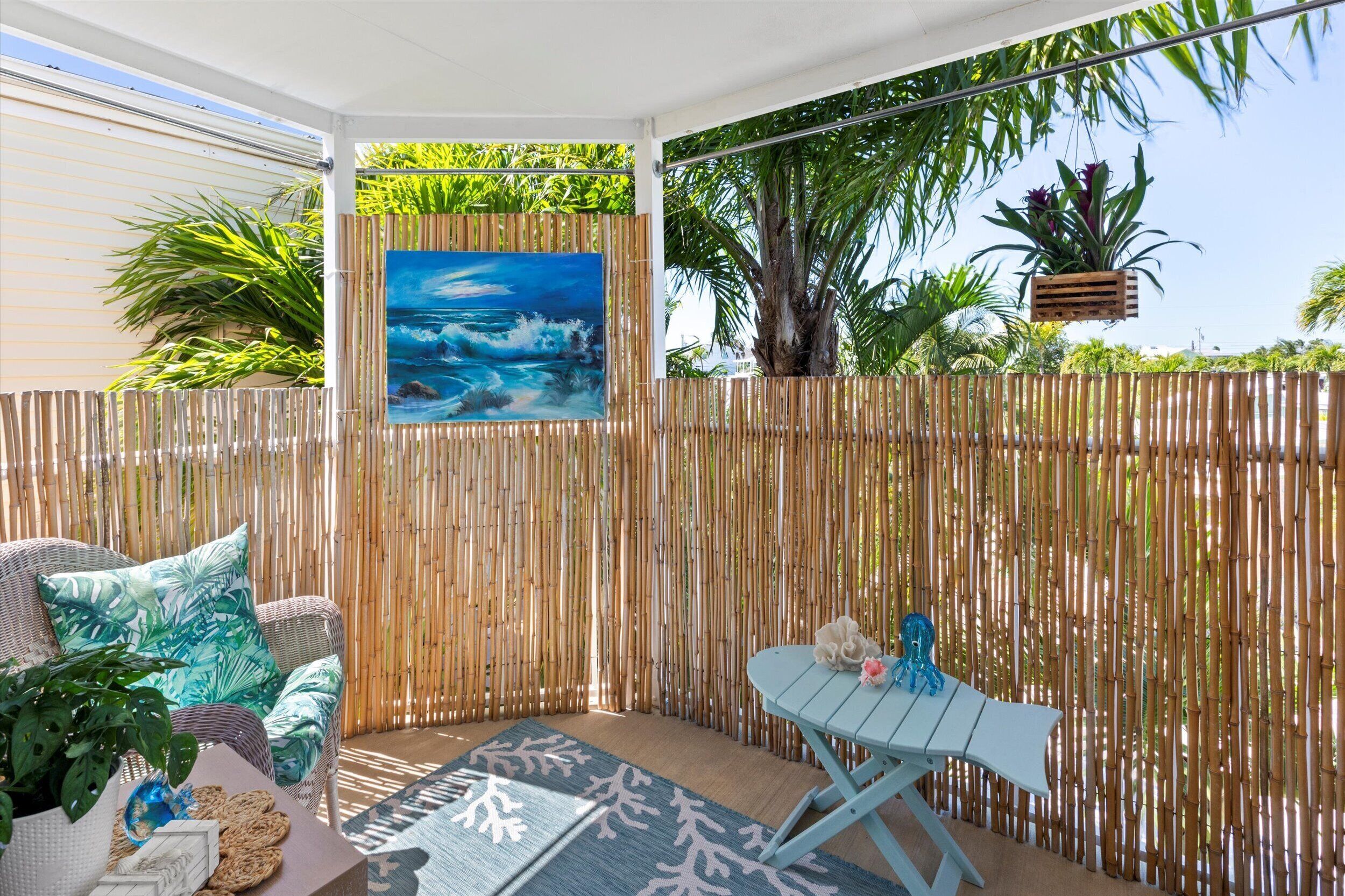 A covered outdoor seating area with a bamboo fence, wicker furniture, and a painting of a beach scene.