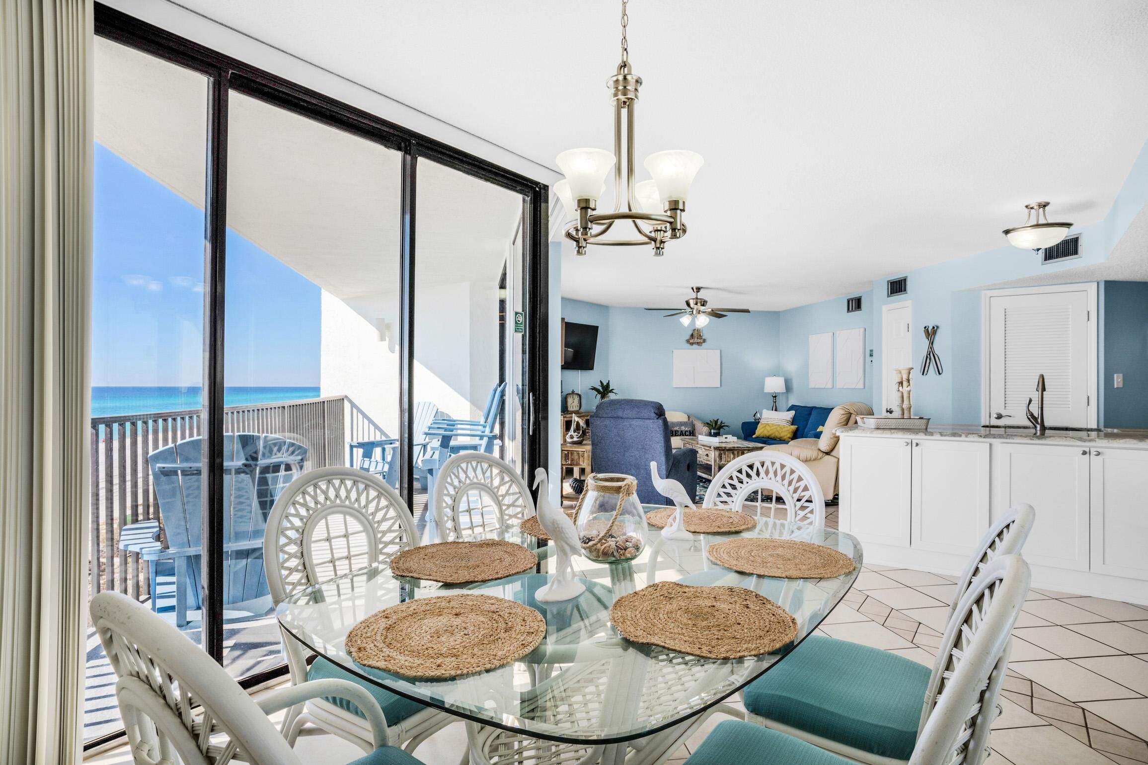 A dining area with a glass table, white chairs, and woven placemats. A chandelier hangs above, and a ceiling fan is visible. The room has a view of the ocean through large glass doors.