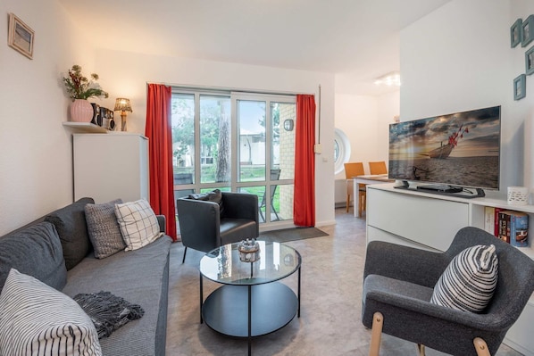 Ein modernes Wohnzimmer mit einer Couch, einem Sessel und einem Couchtisch, sowie ein Essbereich mit Tisch und Stühlen und einem an der Wand montierten Fernseher.