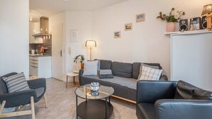 Ein modernes Wohnzimmer mit einer Couch, zwei Sesseln, einem Couchtisch und einem durchgehenden Gang sichtbarem Küchenbereich.