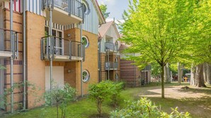 Un edificio residencial con varios balcones, fachada de ladrillo y un pequeño jardín con árboles y arbustos.
