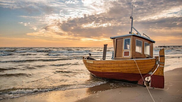 Un bote de madera con cabina, amarrado en una playa arenosa con olas, bajo un cielo parcialmente nublado.