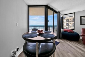 A modern apartment with a balcony overlooking the sea, a round dining table with black mats, and a cozy seating area with a red cushion. - Sunbird 1010e (Panama City Beach)
