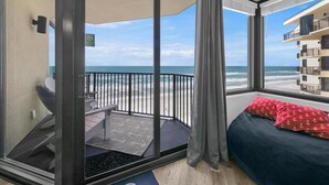 A balcony with a view of the beach, a bed with red pillows, and a small seating area. - Sunbird 1010e (Panama City Beach)