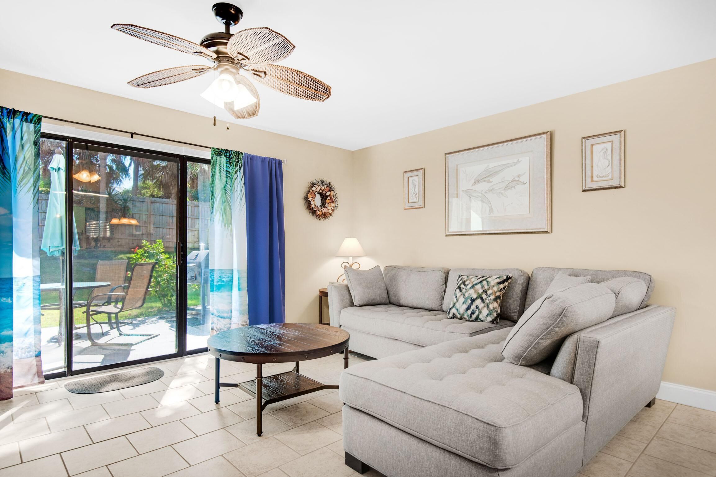 A living room with a grey sectional sofa, a round coffee table, a ceiling fan, and a sliding glass door leading to a patio with outdoor furniture.