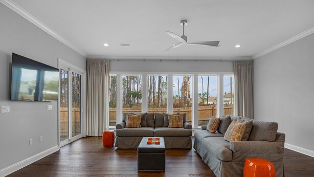 Un amplio salón con suelo de madera, ventilador de techo y grandes ventanales con vistas al exterior.