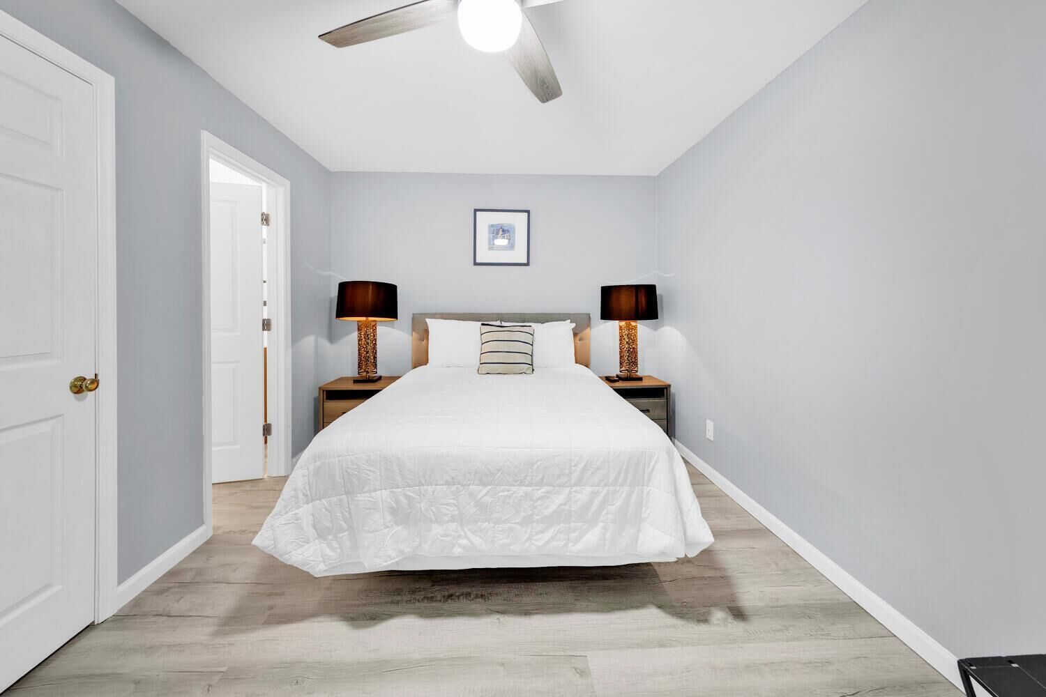 A bedroom with a bed, two bedside tables with lamps, a ceiling fan, and a framed picture on the wall.