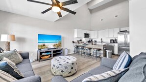 A modern living room with a grey sofa, ottoman, and a ceiling fan. A TV is mounted on the wall, and a kitchen area is visible in the background. - Horizon South 54-303 (Panama City Beach)