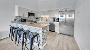 A modern kitchen with a central island, bar stools, stainless steel appliances, and a tiled backsplash. - Horizon South 54-303 (Panama City Beach)