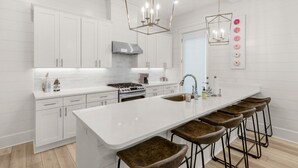 A modern kitchen with white cabinetry, a large island with stools, a stainless steel range hood, and a wall-mounted light fixture.