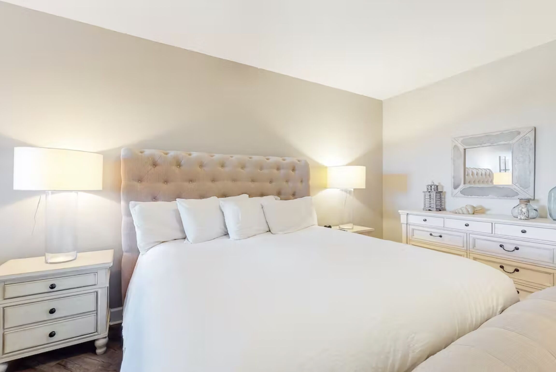 A neatly arranged bedroom with a large bed, two bedside tables with lamps, a dresser, and a mirror on the wall.