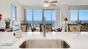 A kitchen with a large window overlooking the sea, a ceiling fan, and a dining area with chairs and a table. - Hidden Dunes 405 (Panama City Beach)