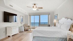A bedroom with a large bed, a ceiling fan, a TV, a dresser, a chair, and a view of the ocean. - Hidden Dunes 405 (Panama City Beach)