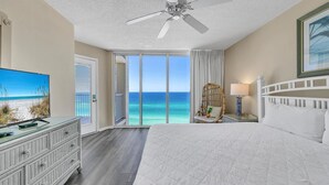 A hotel room with a large bed, a television, a wicker chair, and a view of the ocean.