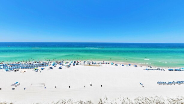 A beach with rows of beach umbrellas and sun loungers, a volleyball net, and a clear view of the ocean.