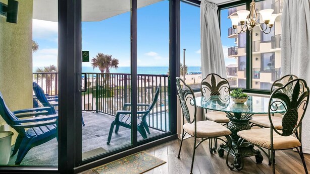 A balcony with a glass door, blue chairs, a glass table with chairs, a chandelier, and a view of the sea and buildings. - Sunbird 211e (Panama City Beach)