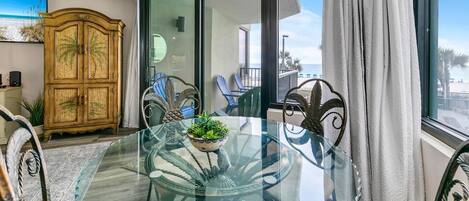 A dining area with a glass table, chairs, a wooden cabinet, and a view of the beach.