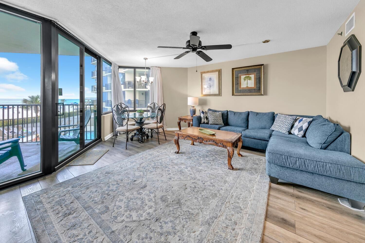 A spacious living room with a blue sofa, a wooden coffee table, and a dining area with a glass table and chairs. There is a balcony with a view of the ocean.