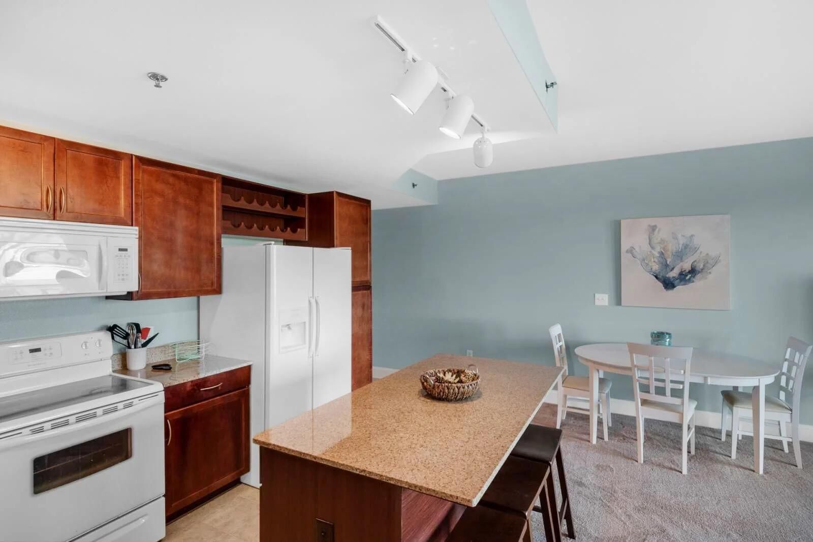 A modern kitchen with wooden cabinets, a white refrigerator, a microwave, and a stove. There is a granite countertop with a bowl and a dining area with a table and chairs.