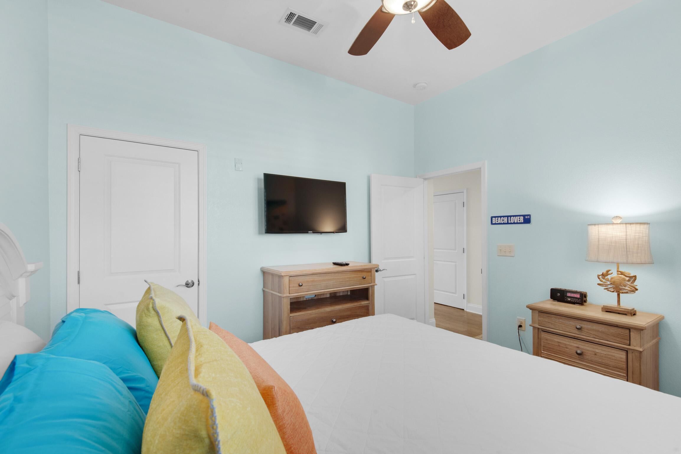 A bedroom with a bed, a television, a ceiling fan, a nightstand, and a sign that reads "BEACH LOVER".