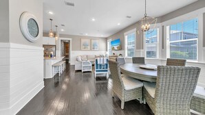 Une cuisine moderne avec un coin repas, comprenant une table ronde et des chaises en osier, un plancher en bois et de grandes fenêtres équipées de stores.