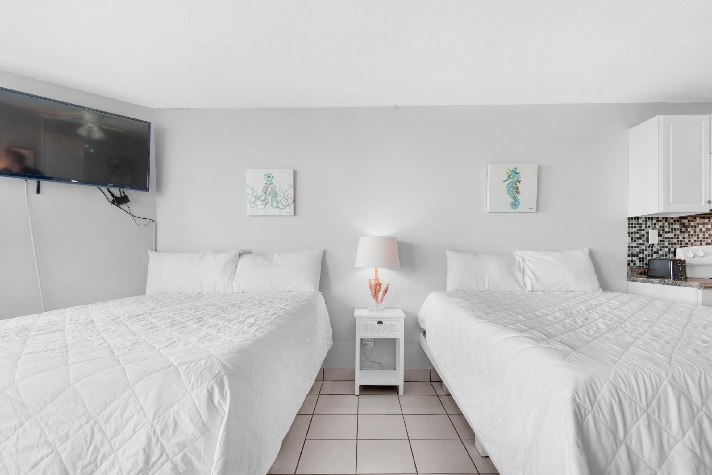 A hotel room with two beds, a TV mounted on the wall, a small table with a lamp, and artwork on the walls.