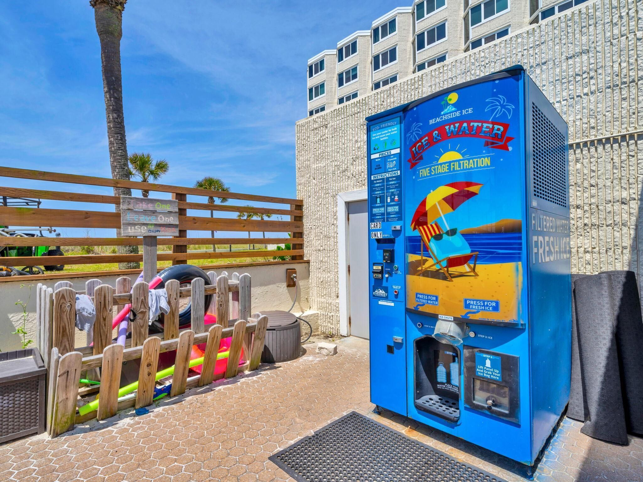 Ein Automatikgerät am Strand, das Eis und Wasser verkauft und über ein fünffach-Filtersystem verfügt, in der Nähe eines Holzzauns und Palmen gelegen.