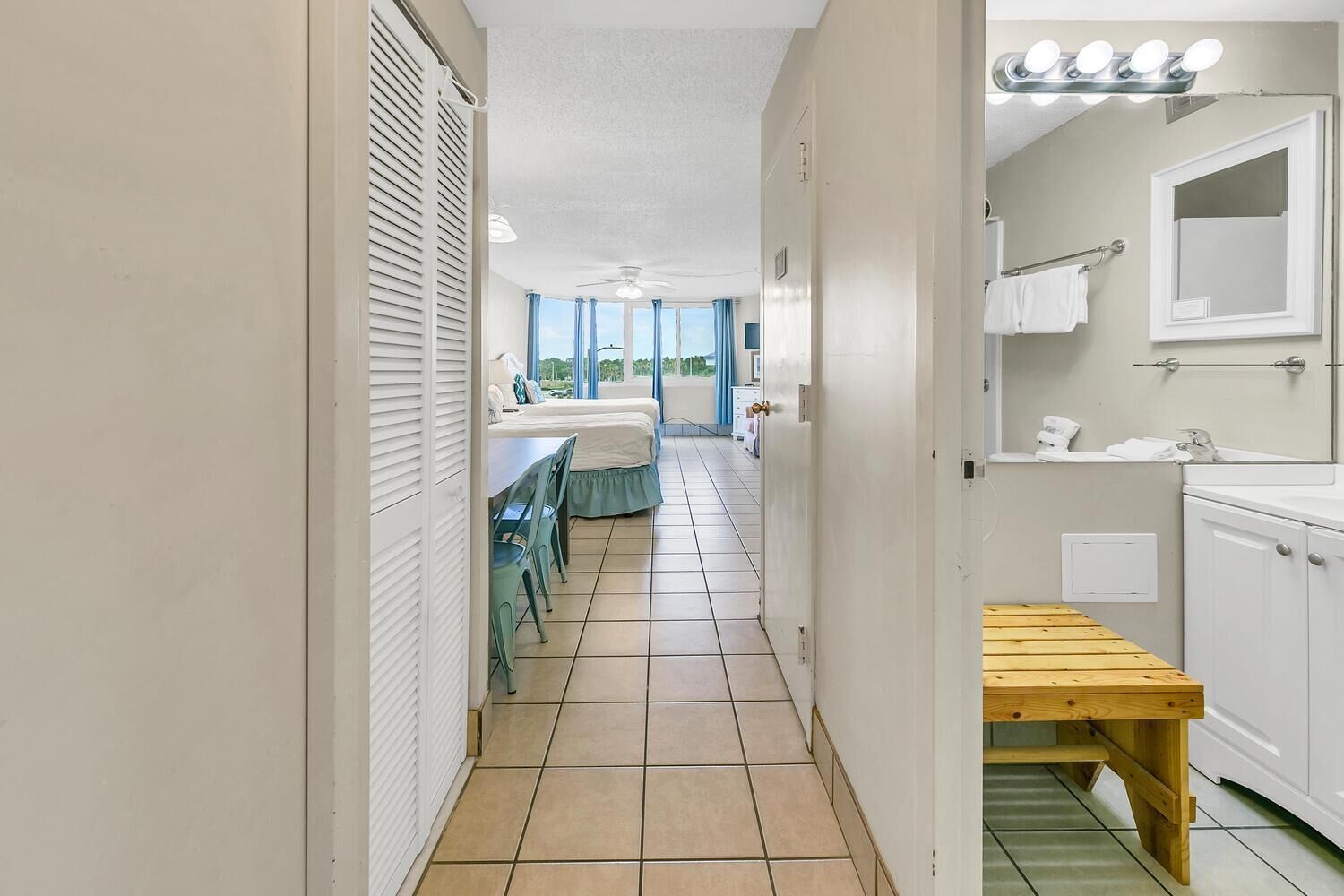 A narrow hallway leading to a bedroom with a bed, a small table, and chairs. The room has a window with curtains, a ceiling fan, and a bathroom with a sink and mirror.