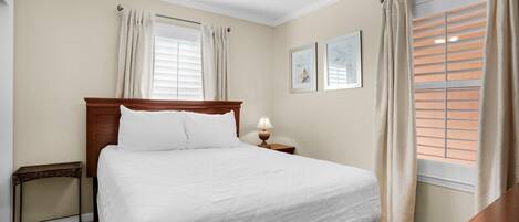 Een net opgemaakt bed in een hotelkamer, met een houten hoofdbord, een nachtkastje met een lamp en een raam met zonwering.