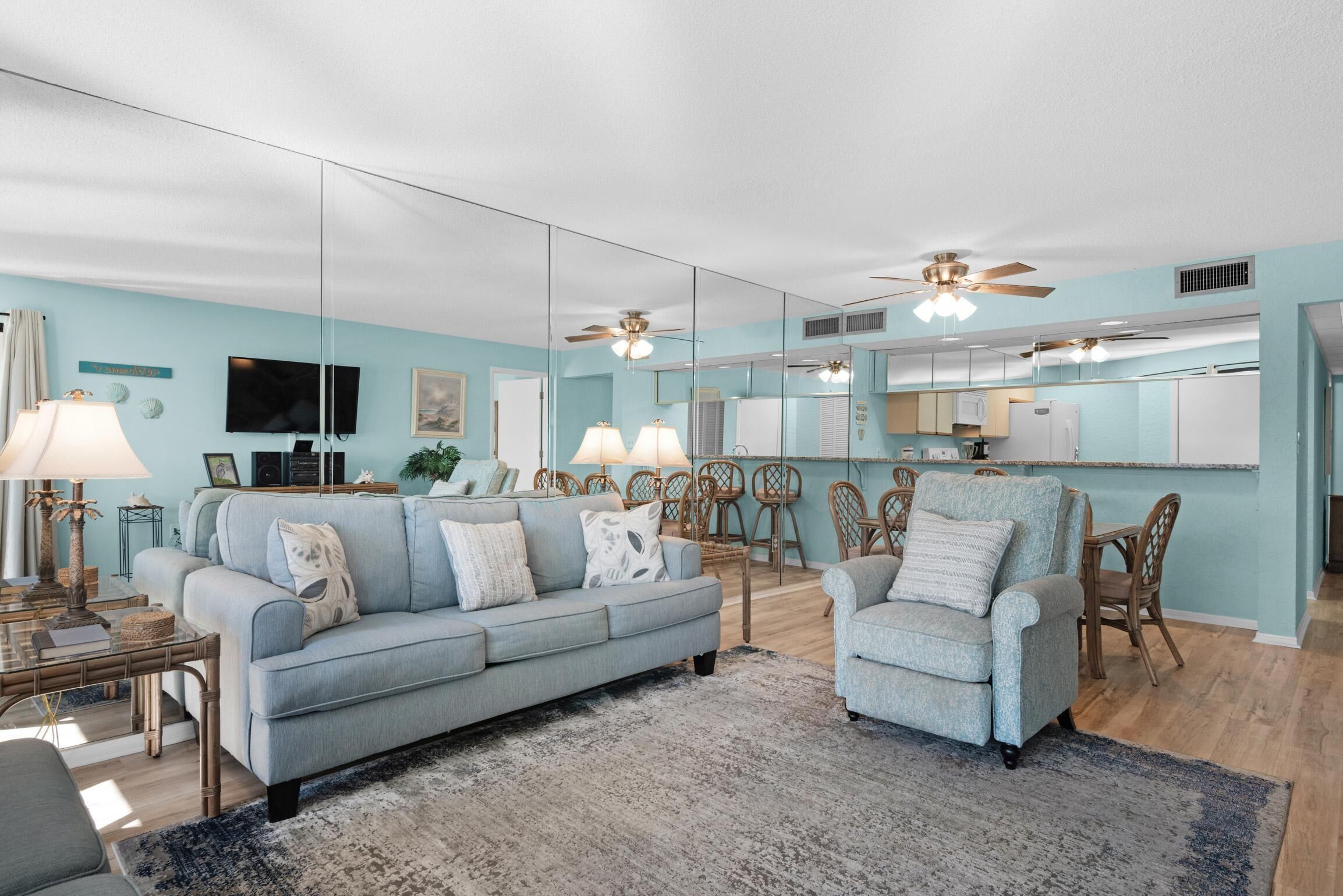 Amplia sala de estar con un sofá azul claro, un sillón a juego y un comedor con sillas de mimbre. Un espejo grande refleja la habitación, y se observan ventiladores de techo.