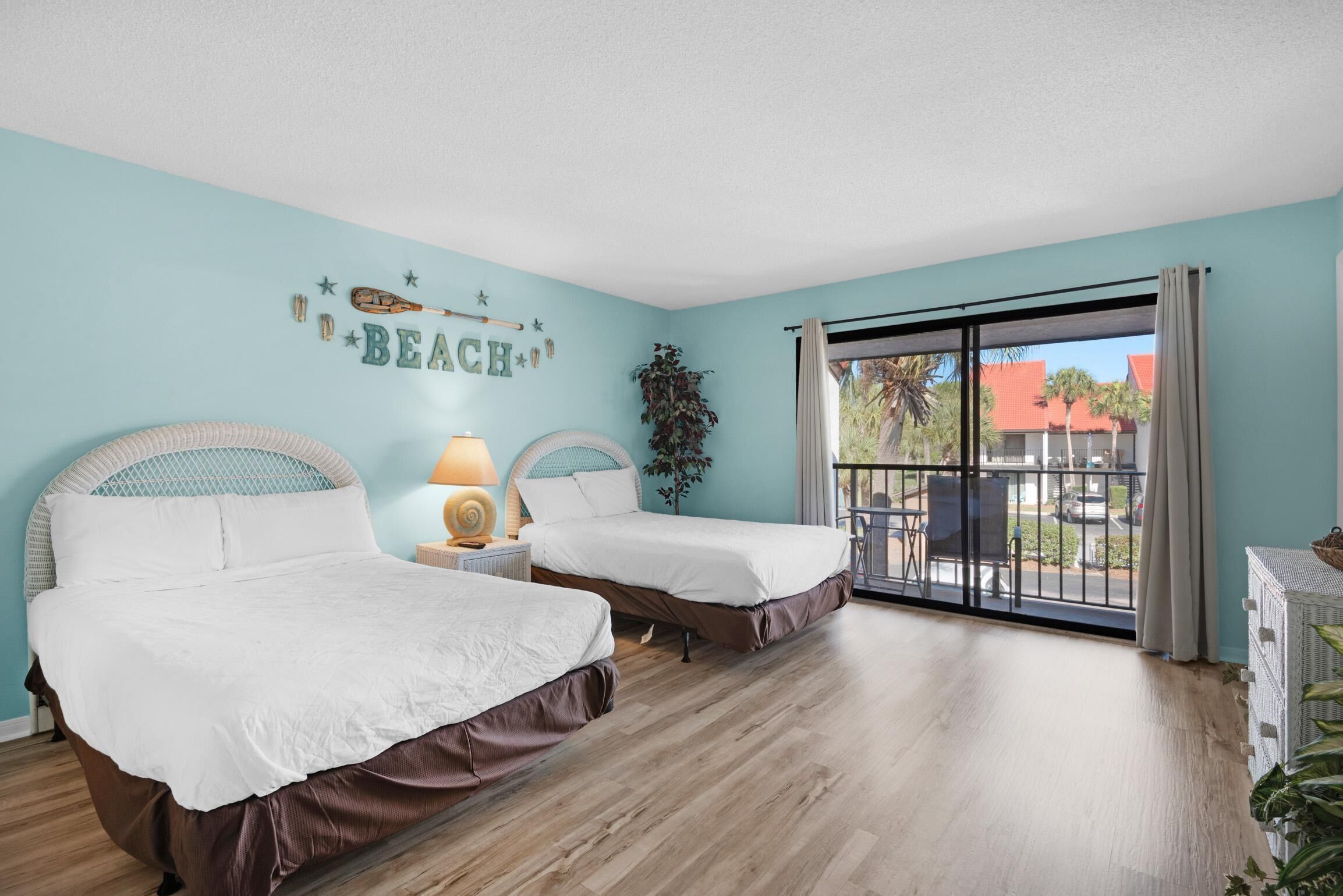 A room with two beds, a nightstand with a lamp, a wall decoration, and a view of the outside.