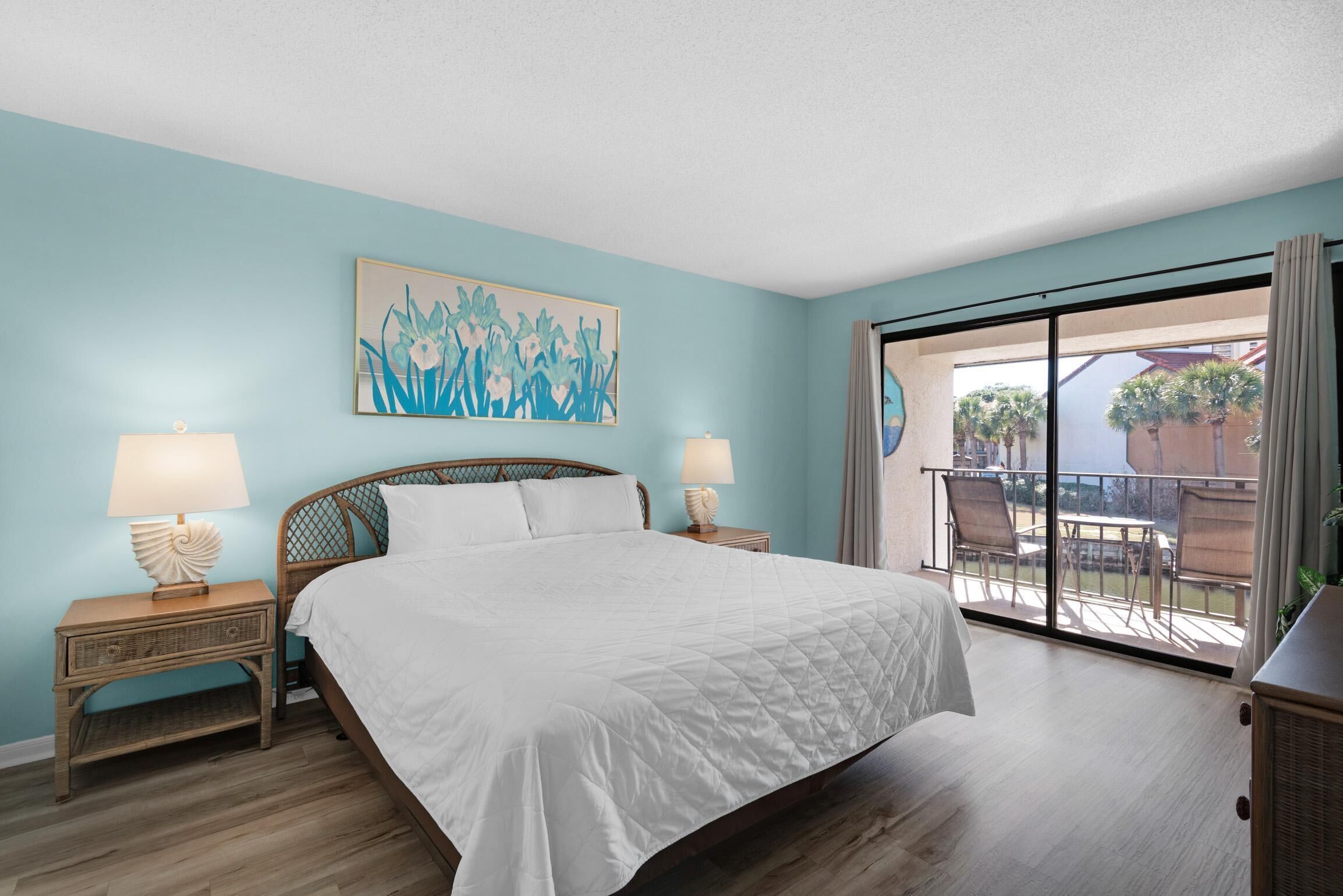 A bedroom with a bed, two bedside tables with lamps, a large window, and a balcony with outdoor furniture.