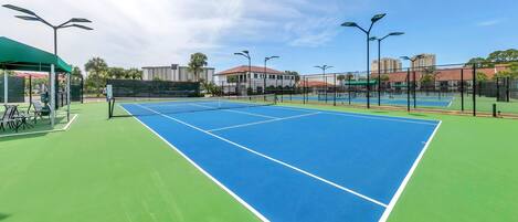 A tennis court with green and blue surfaces, surrounded by a fence, and a covered seating area with chairs.