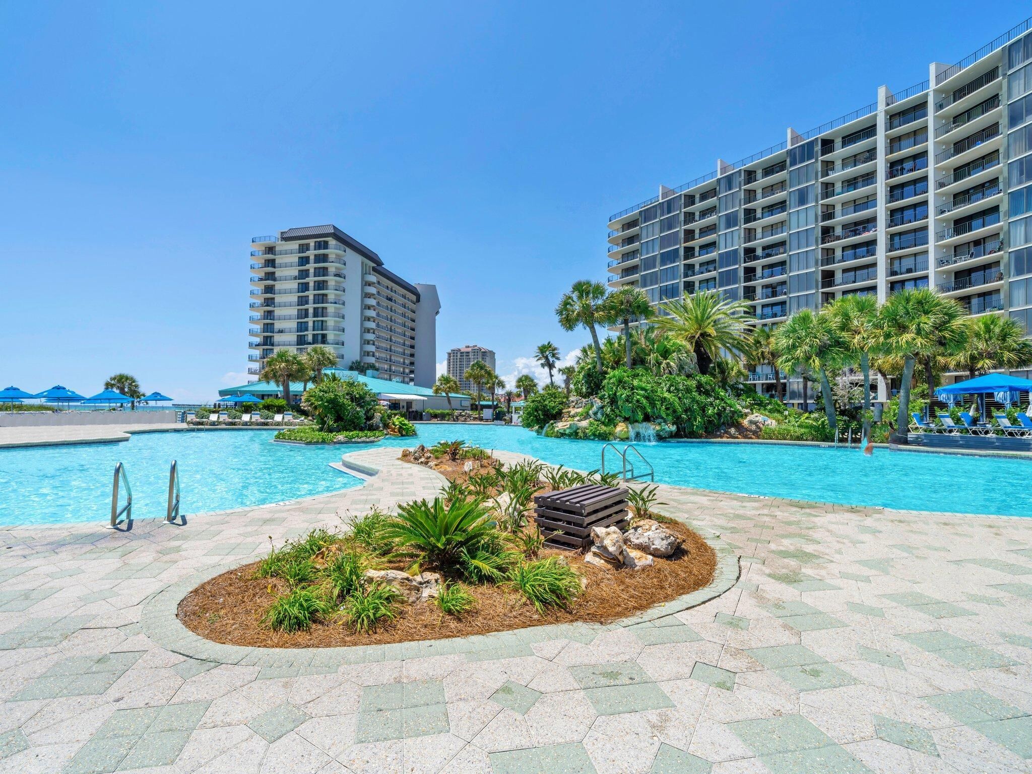 A hotel pool area with a clear blue pool, surrounded by palm trees and lounge chairs.