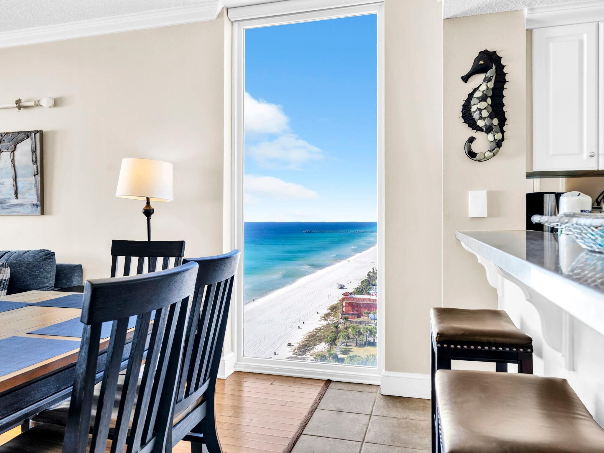 A room with a view of a beach and ocean, a dining area with a table and chairs, and a kitchen area with a seahorse decoration.
