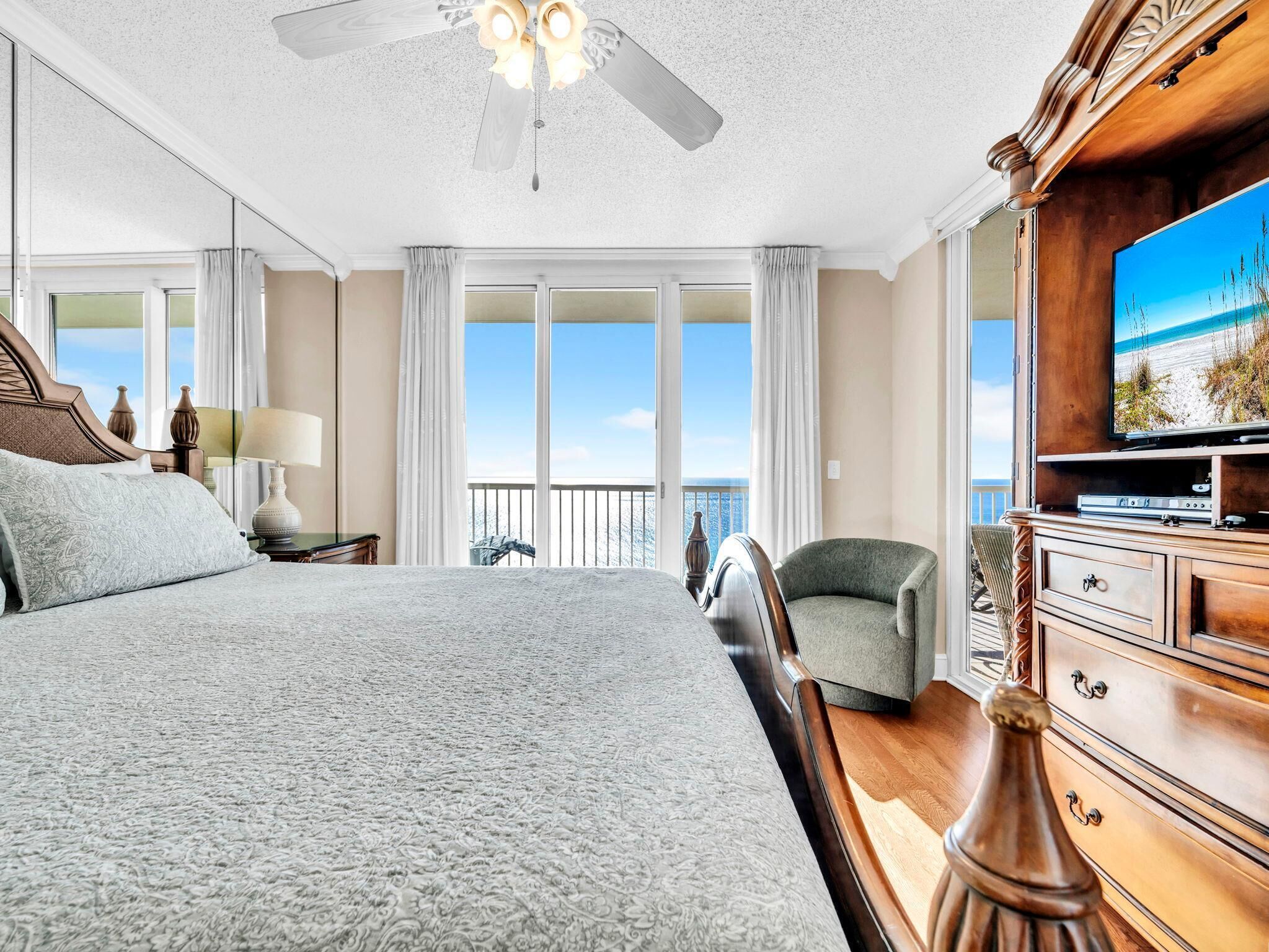 A hotel room with a large bed, a television, a chair, and a balcony with a view of the beach.