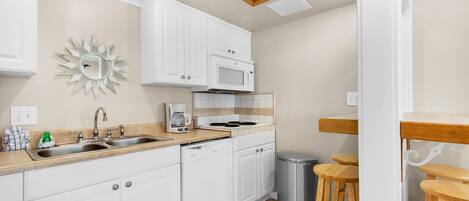A modern kitchen with white cabinetry, a stainless steel sink, a microwave, and a coffee maker. There is a trash can and wooden stools in the corner.