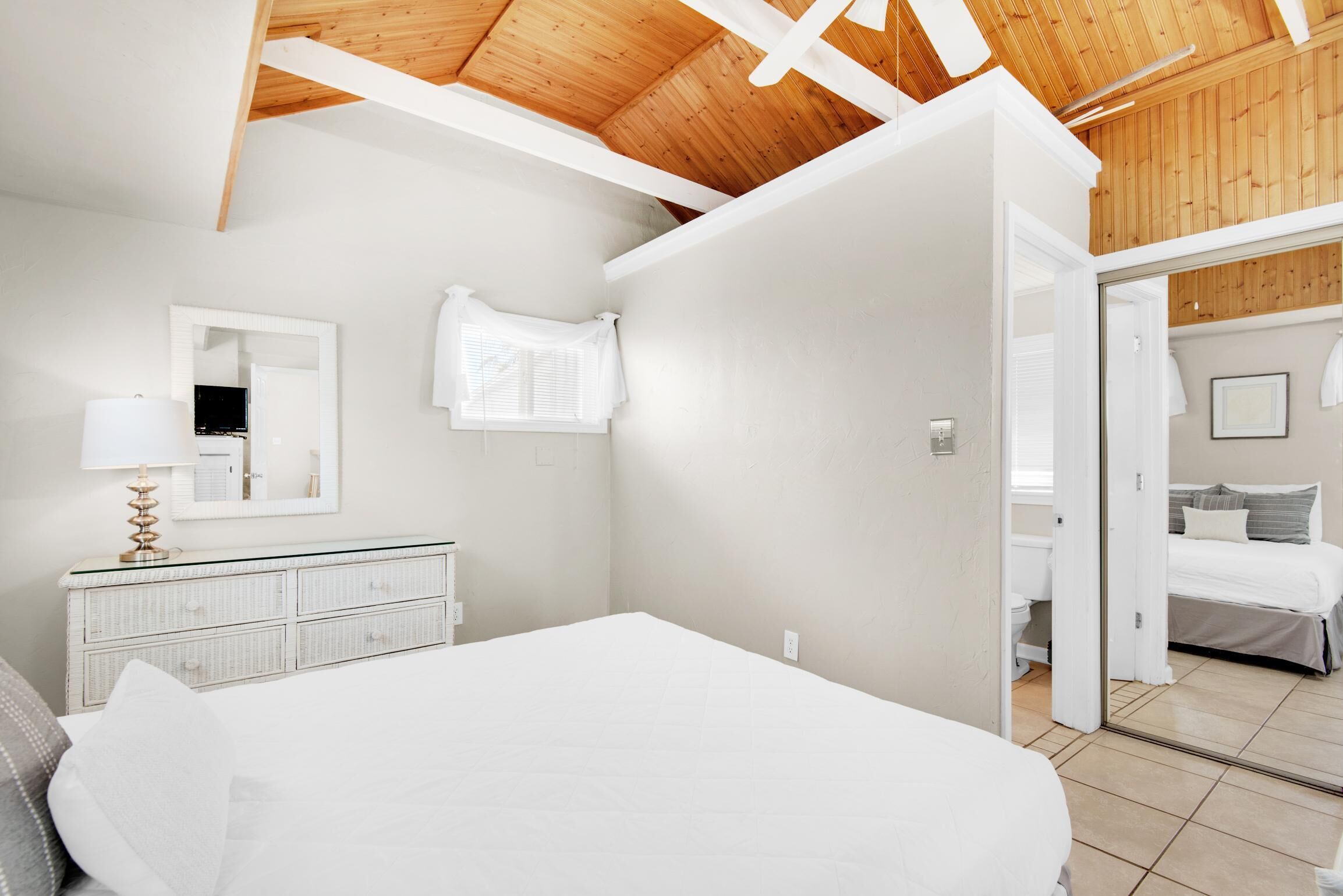 A bedroom with a wooden ceiling, a white bed, a dresser with a mirror, and a door leading to another room.