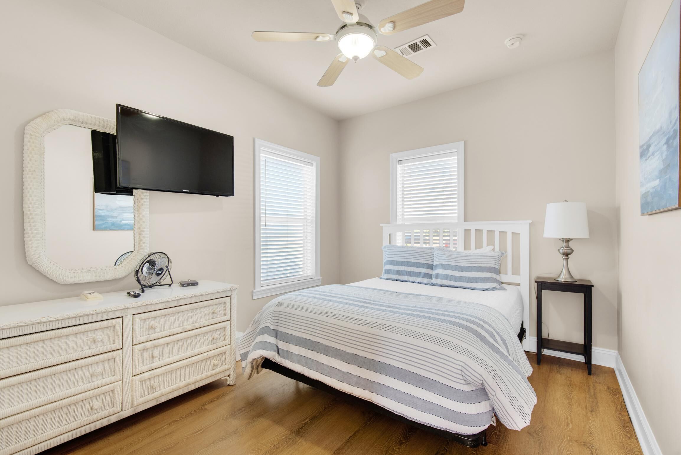 A bedroom with a bed, dresser, TV, ceiling fan, windows, and a lamp.